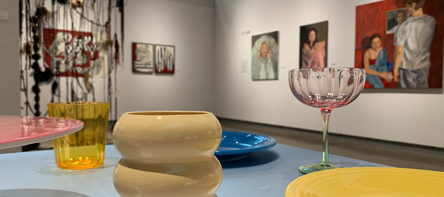 colorful glasses and serving dishes arranged on a blue silk table cloth, in front of several portrait paintings and some abstract paintings; also nearby is an installation made of many lengths of braided hair draped from a dowel suspended from the ceiling