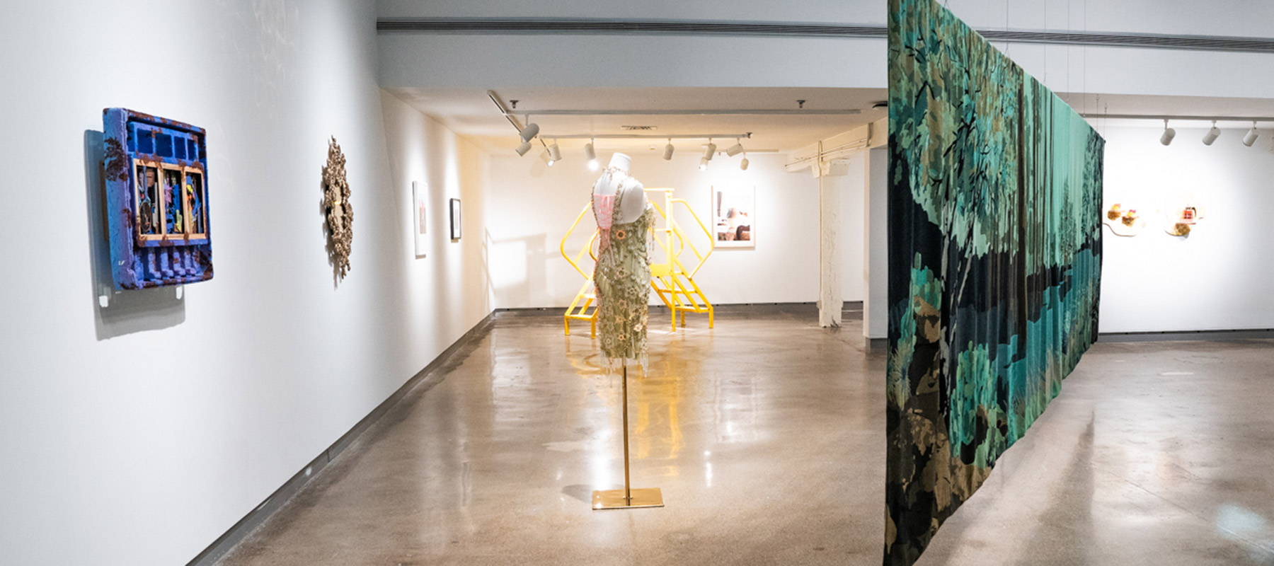 gallery space displaying artwork, including a large tapestry depicting a lush green landscape, several artworks on the wall, and a dress made of gold chains and pieces of acrylic displayed on a mannequin