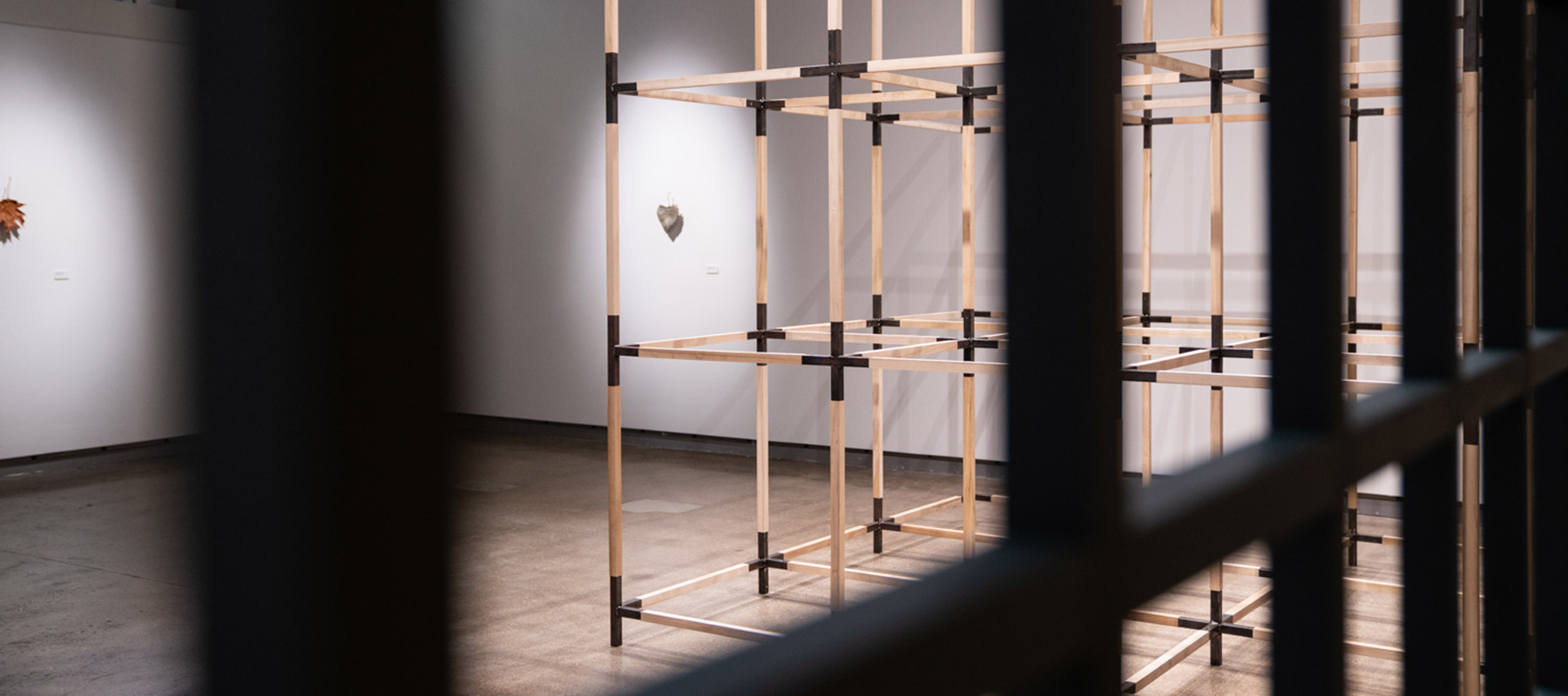 A large, free-standing, three-dimensional grid constructed of wood beams and metal brackets at each intersection, with two carved wooden leaves mounted on the wall behind it. The artwork is viewed through a flat, upright grid structure.