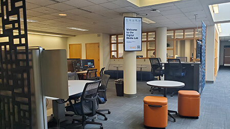 A photo of the Digital Media Lab lobby, including tables, chairs, orange cushion seats, and a digital sign mounted on a pillar