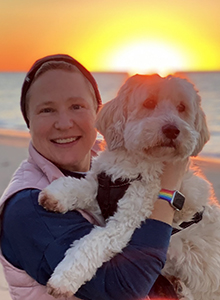A woman smiling while holding her dog in front of sunset