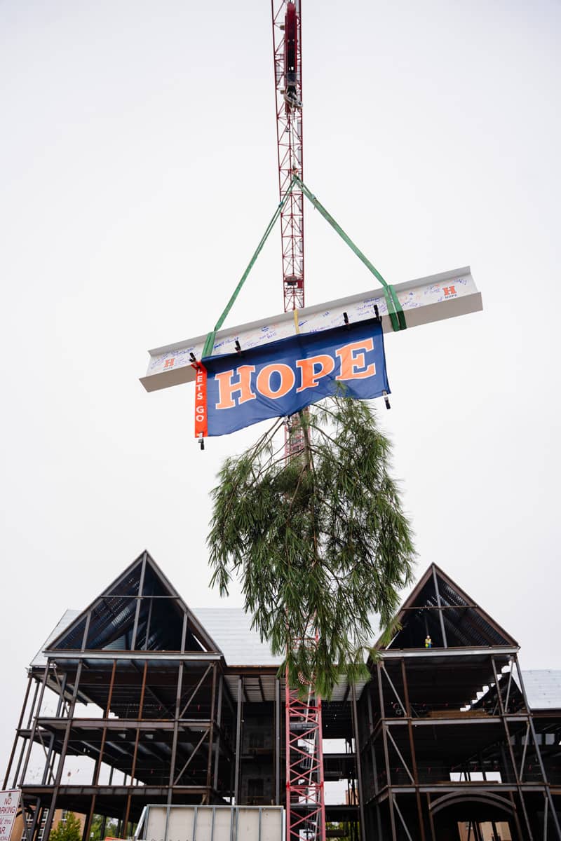 The final beam being lifted into place by a crane for the Economics & Business building topping off ceremony. A HOPE flag and an evergreen tree limb are hanging from the beam.
