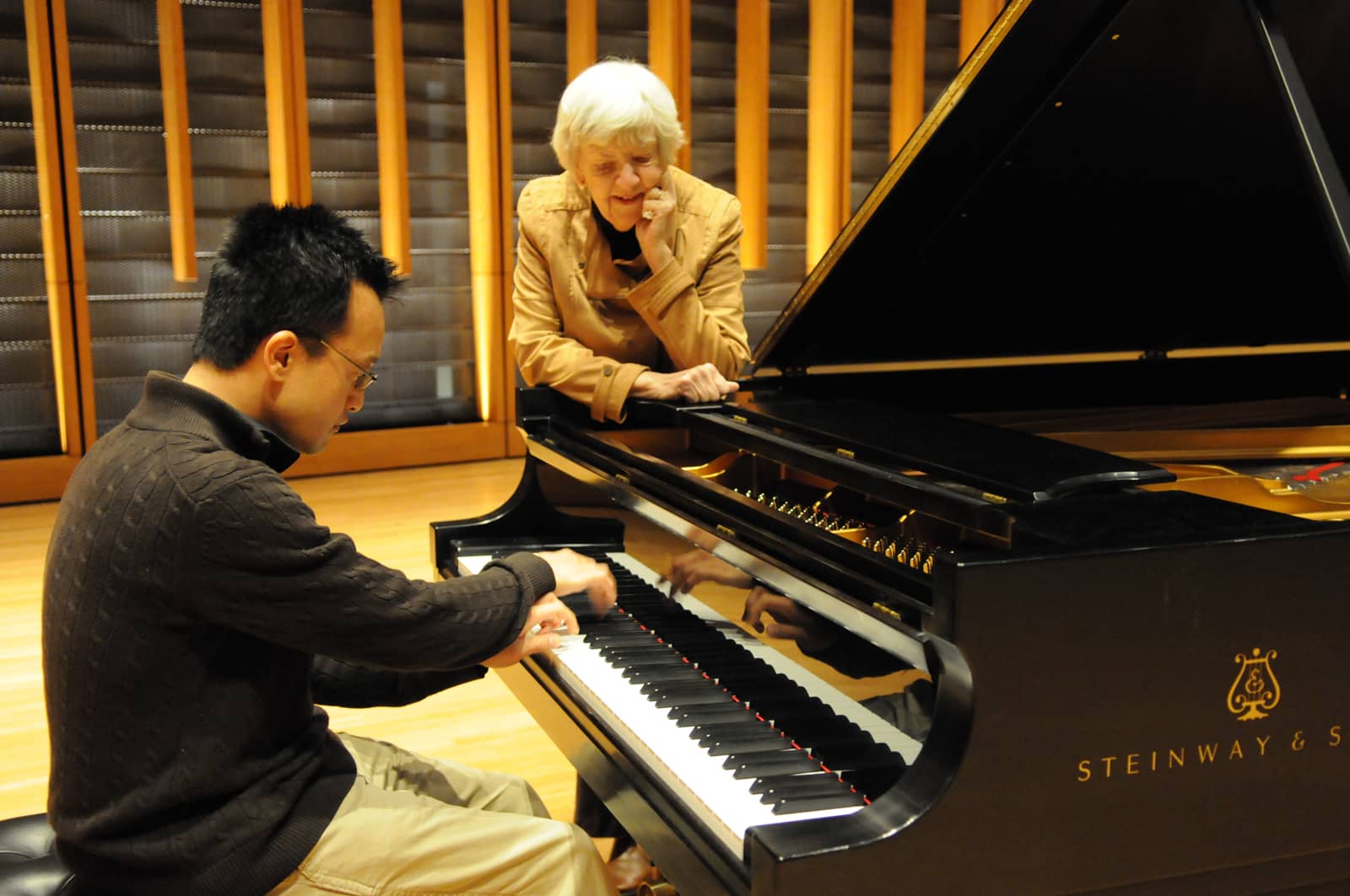 Professor Joan Conway standing at a Steinway grand piano, instructing the man who is playing